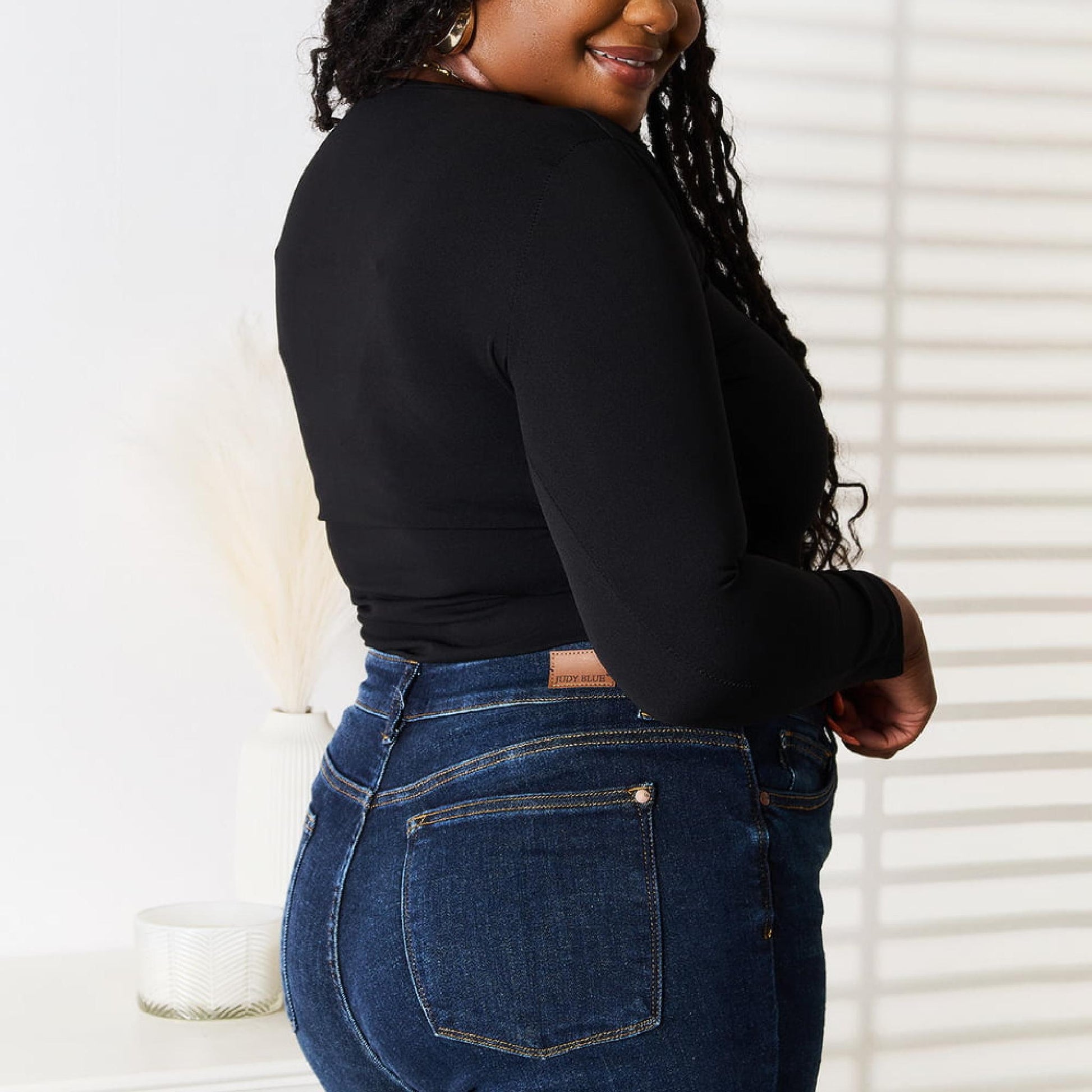 Woman wearing a black long-sleeve top and blue jeans standing in a room with white walls and furniture.