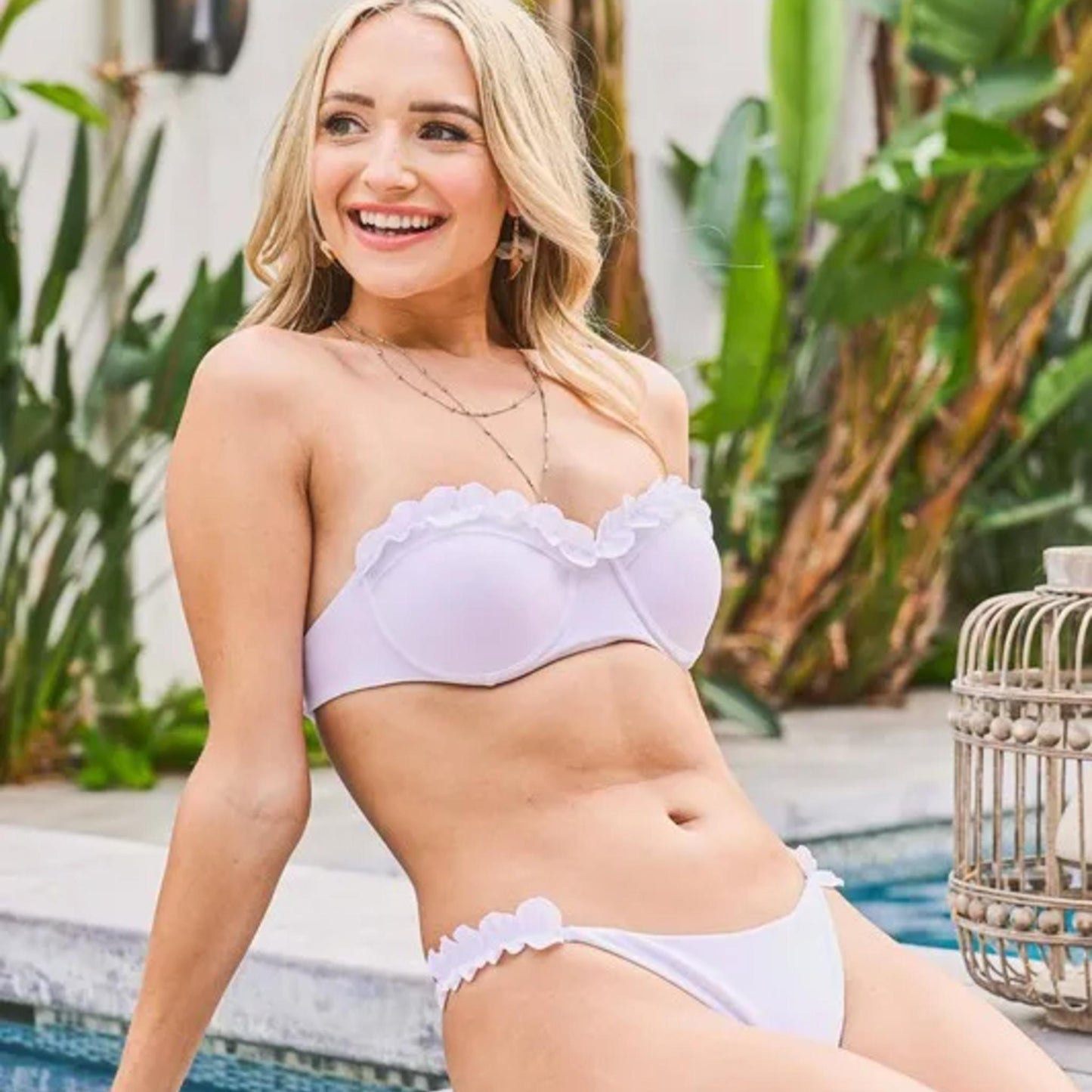 Woman in a white bikini sitting by a pool with plants in the background