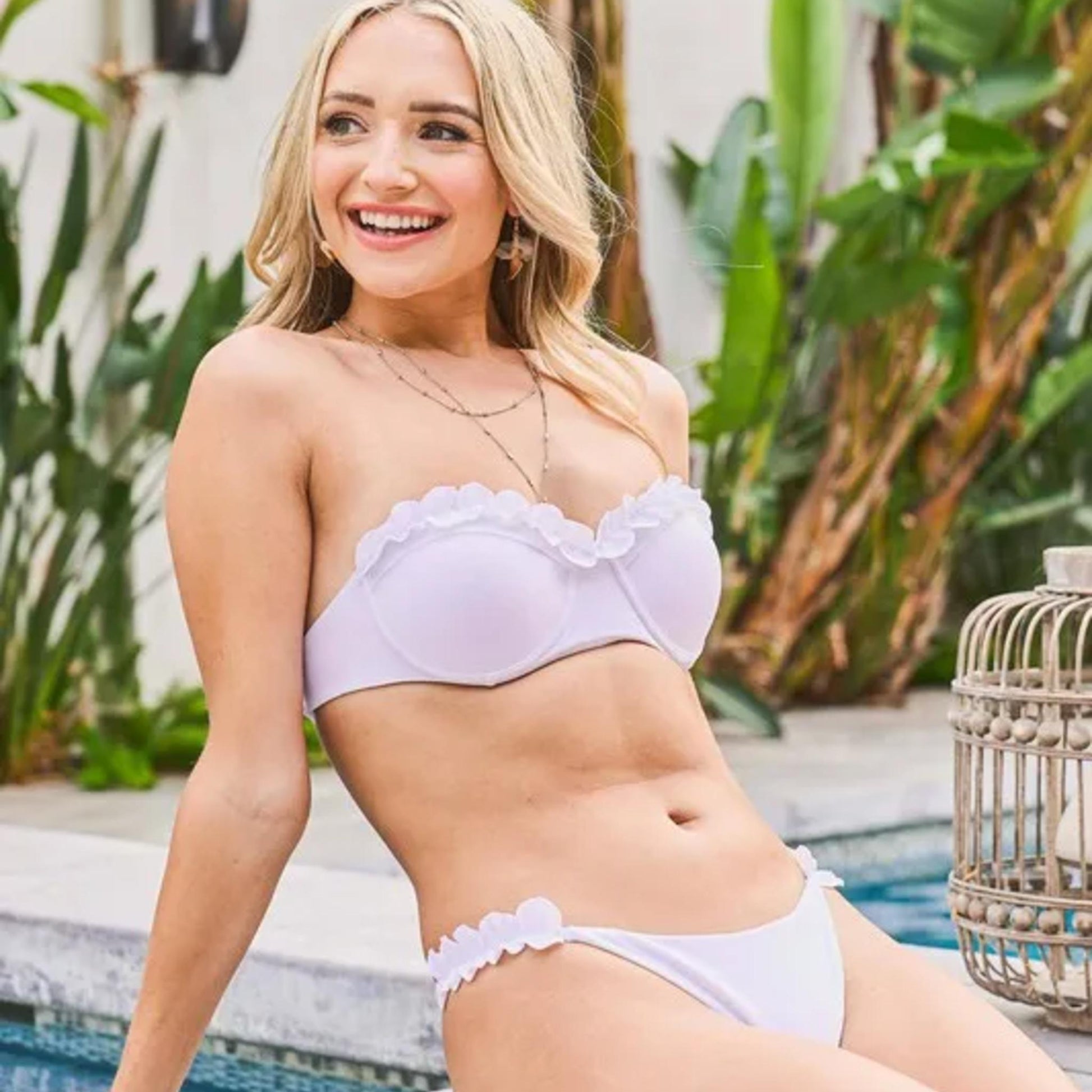 Woman in a white bikini sitting by a pool with plants in the background