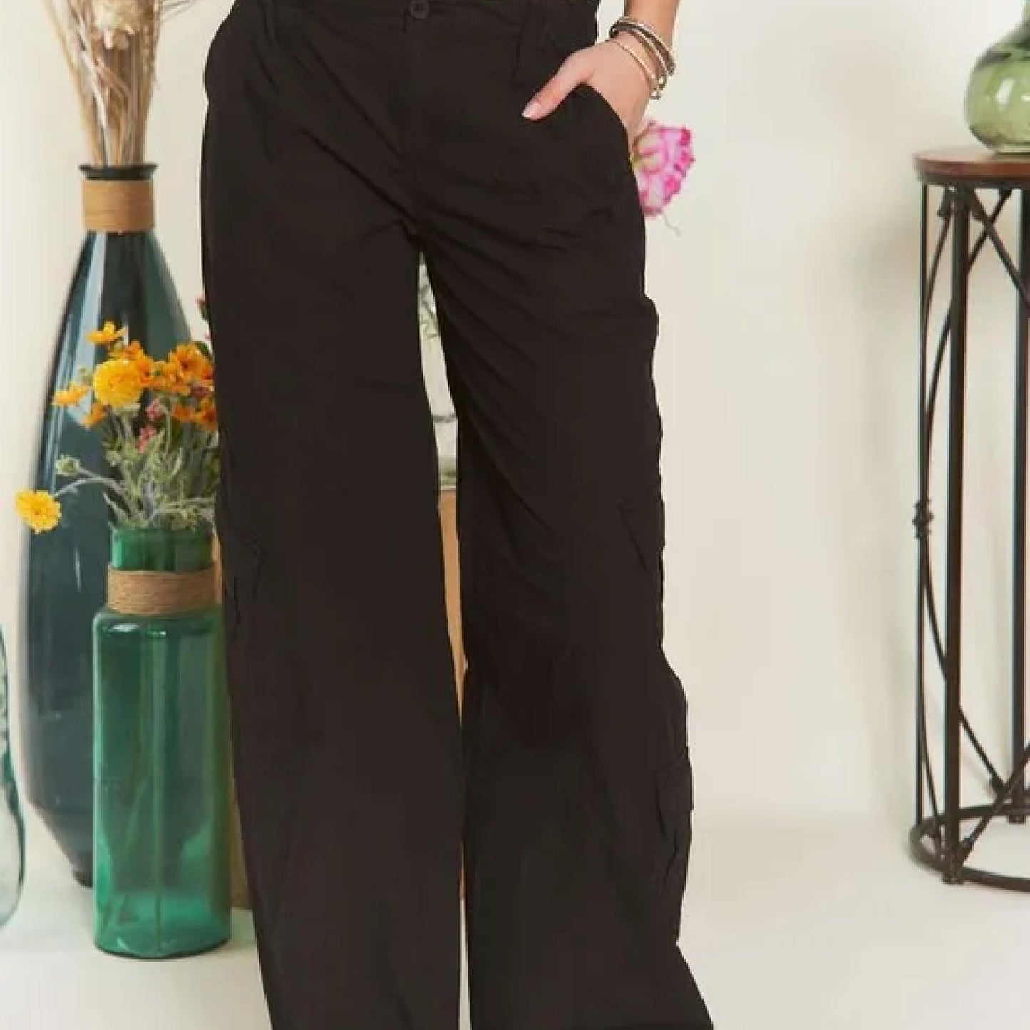 Person wearing black wide-leg pants standing in a room with decorative vases and flowers.