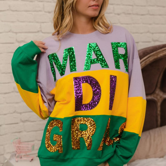 Person wearing a colorful 'MARDI GRAS' sweatshirt with sequin letters against a brick wall.