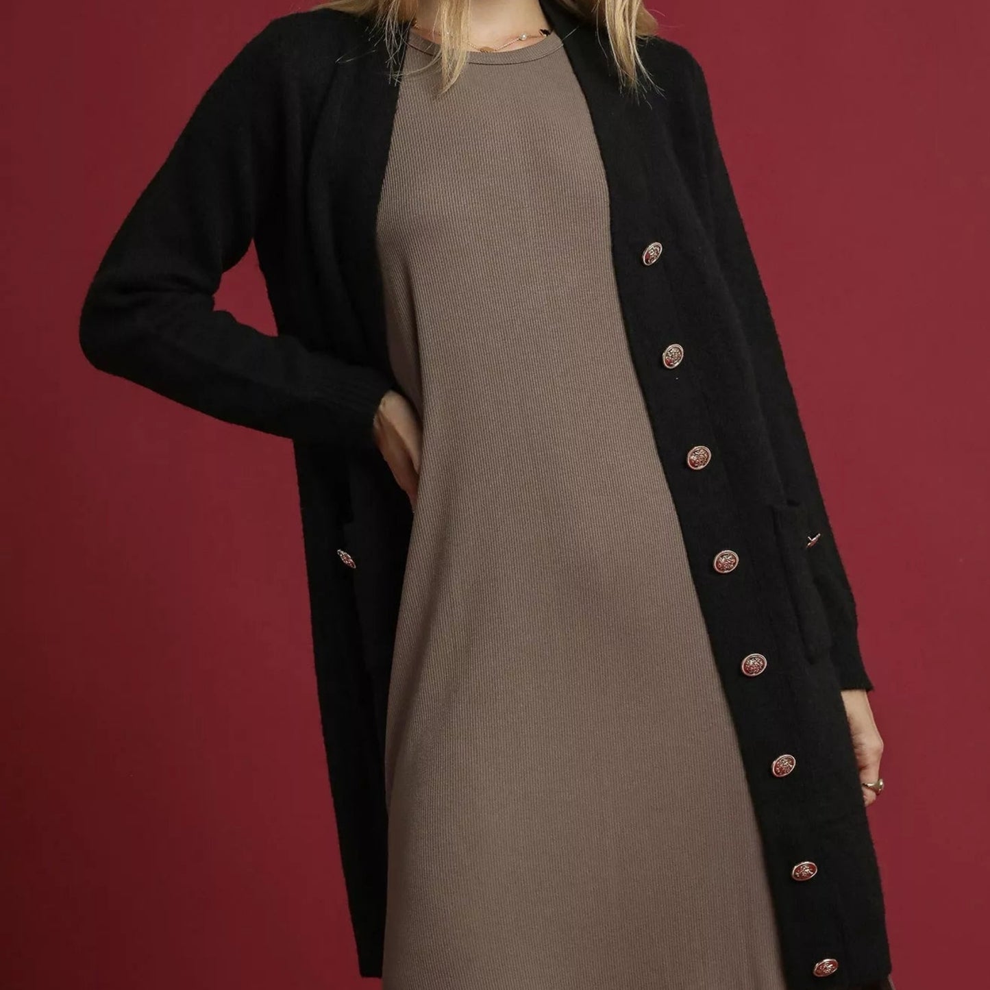 Person wearing a black coat with gold buttons over a brown dress on a red background