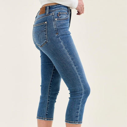 Woman wearing light blue denim capris and a white long sleeve top standing in a bright room.