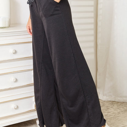 Person wearing dark gray wide-leg pants in a room with a white dresser and curtains.